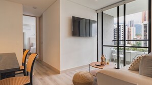 Ein modernes Wohnzimmer mit Sofa, Esstisch und Stühlen, einem an der Wand befestigten Fernseher und einem großen Fenster mit Blick auf die Stadt.