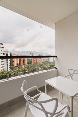 Ein Balkon mit weißem Tisch und Stuhl, mit Blick auf eine Stadtlandschaft mit Gebäuden und Grünflächen.