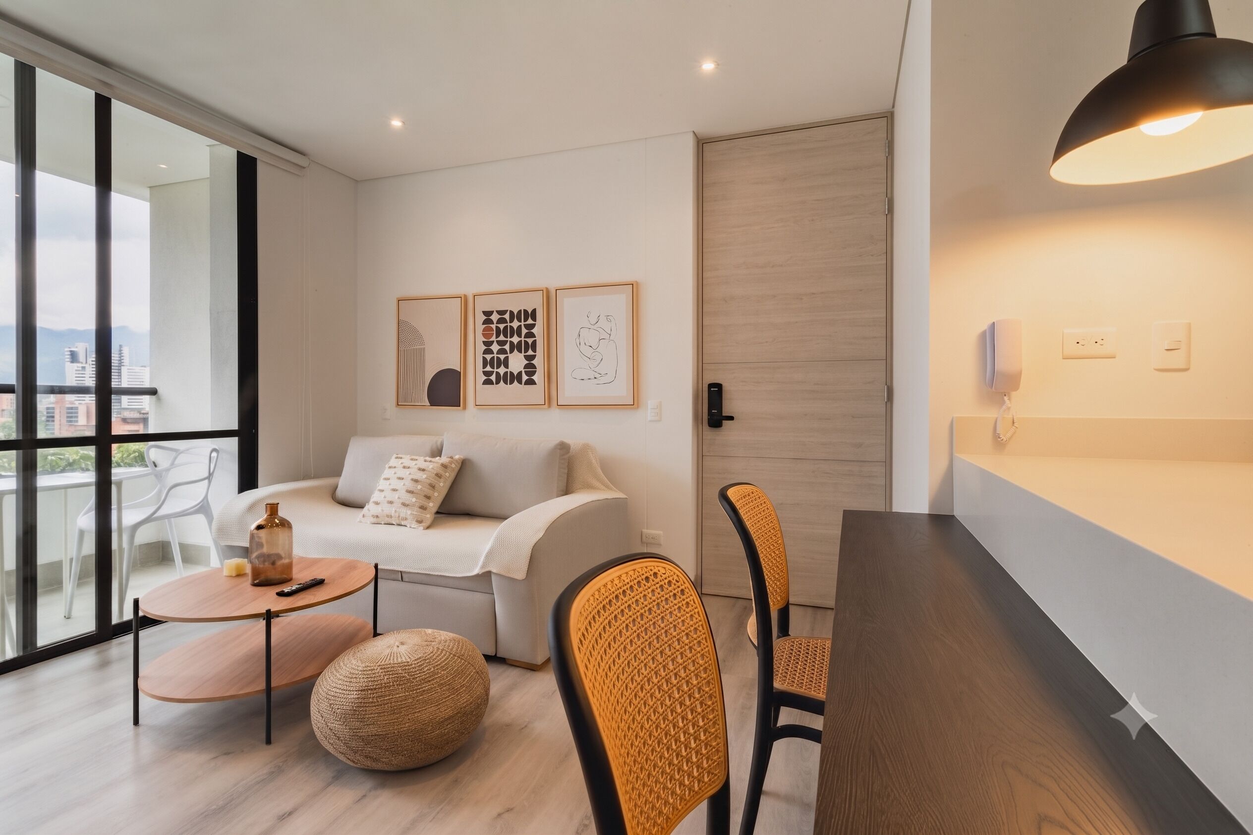 Un salon moderne avec un canapé, une table basse et un fauteuil. On peut y admirer des œuvres d’art encadrées au mur, une porte en bois et une fenêtre donnant sur un paysage.
