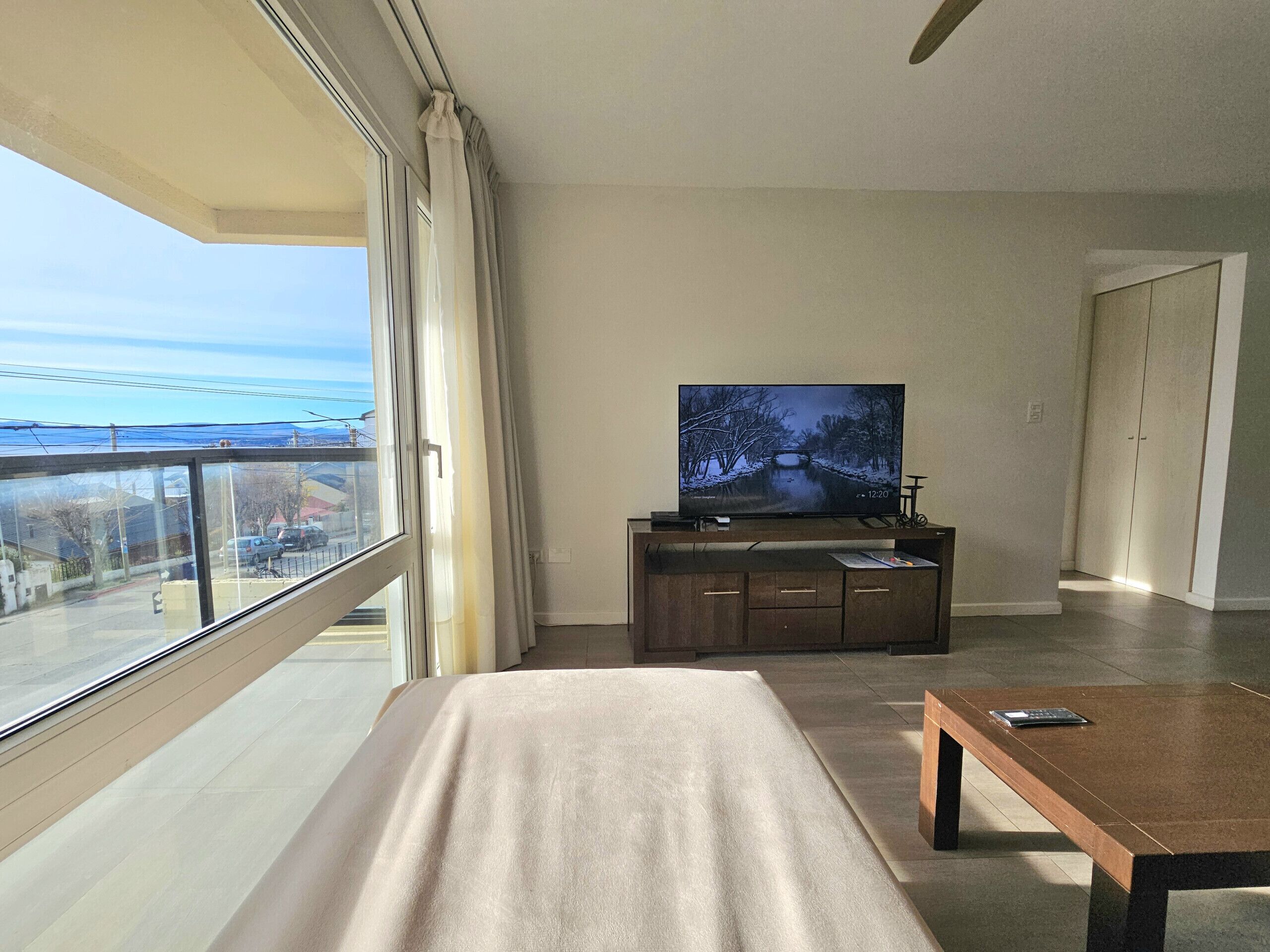 A modern bedroom with a large window, a bed, a TV on a wooden stand, and a balcony.