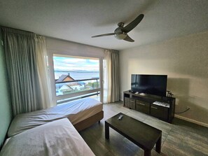 Ein Hotelzimmer mit Bett, Fernseher auf einem Schrank, Deckenventilator und Meerblick durch ein Fenster.