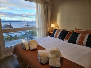 A hotel room with a large window, a bed with multiple pillows, a nightstand with a lamp, and towels on the bed. - Araucaria with LAKE views and parking (San Carlos de Bariloche)