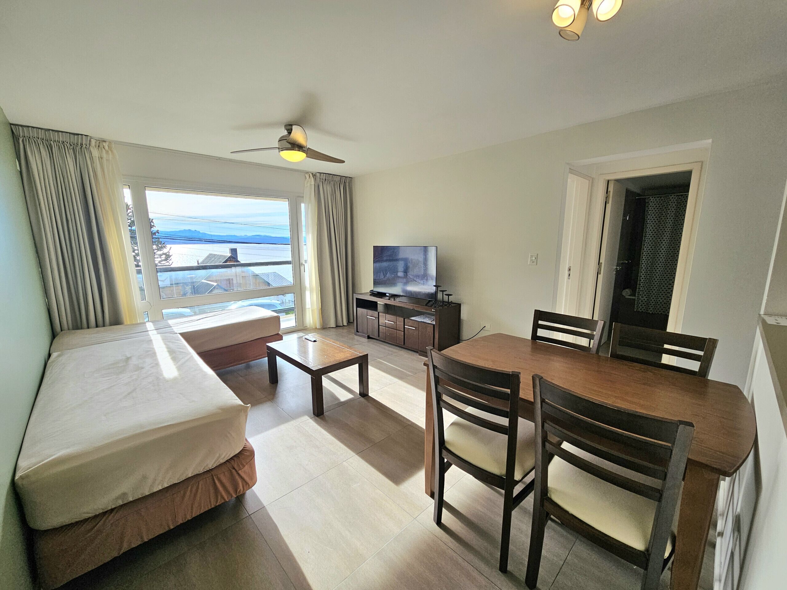 A hotel room with a bed, a desk, a dining table with chairs, a television, and a view of the outdoors.