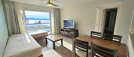A hotel room with a bed, a desk, a dining table with chairs, a television, and a view of the outdoors.