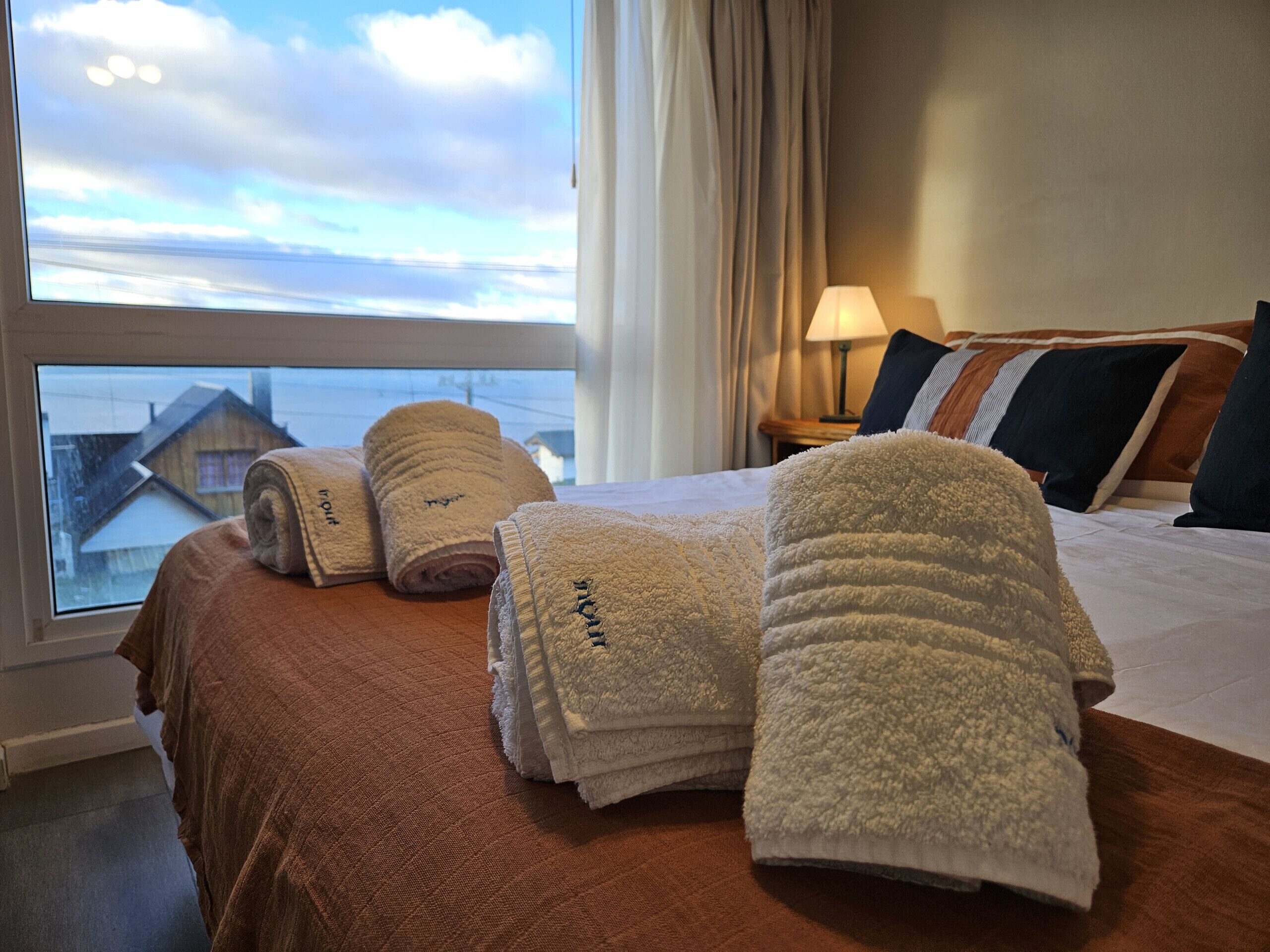 A hotel room with a bed, a window with a view of houses, and several towels on the bed.