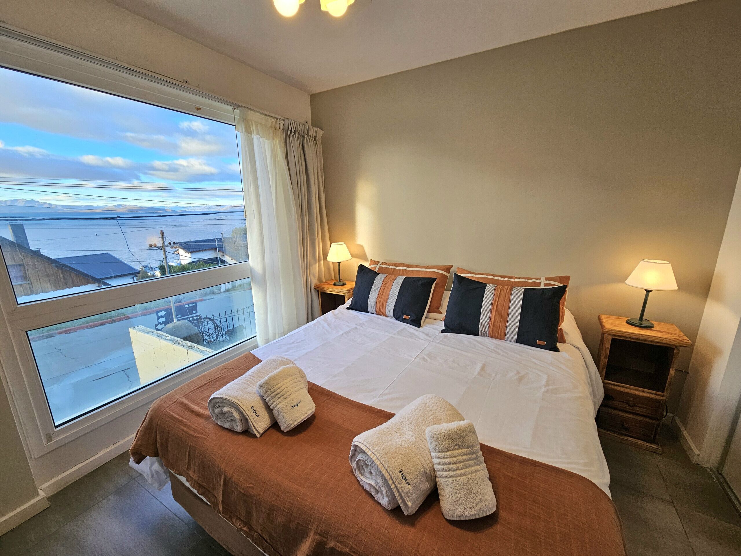 A hotel room with a large bed, two bedside tables with lamps, a window with a view of the sea, and towels on the bed.