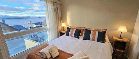 A hotel room with a large bed, two bedside tables with lamps, a window with a view of the sea, and towels on the bed.