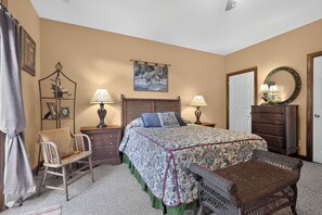 Ein Schlafzimmer mit einem Bett, zwei Nachttischen, einer Kommode, einem Sessel, einer Bank, einer Tür und einem an der Wand befestigten Spiegel.