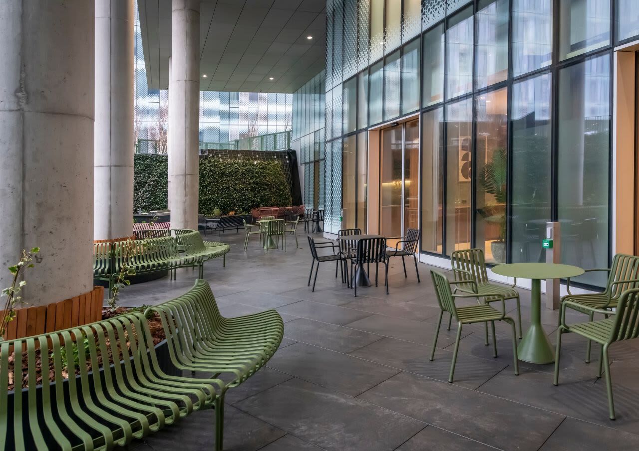 An indoor seating area with green chairs and tables, large windows, and a green wall with plants.