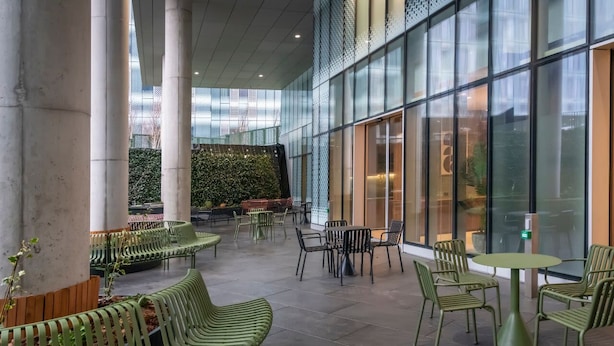 An indoor seating area with green chairs and tables, large windows, and a green wall with plants. - Student Only Square Gardens Acer (Manchester)