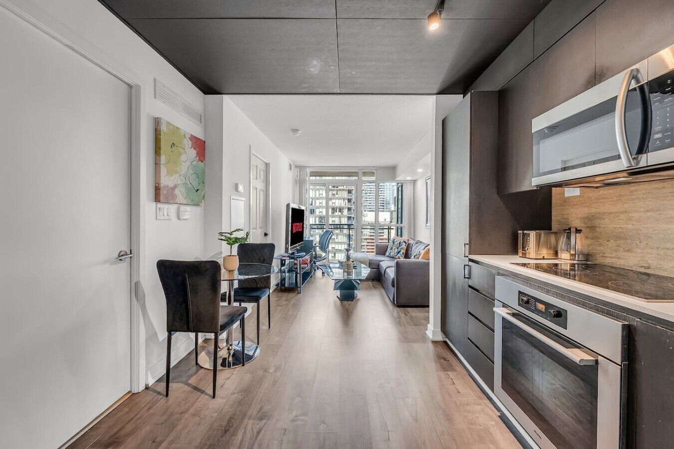A modern kitchen with a built-in oven and microwave, a dining area with a glass table and chairs, and a living room with a sofa and a TV.