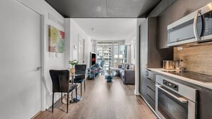 Eine moderne Küche mit eingebautem Ofen und Mikrowelle, ein Essbereich mit Glastisch und Stühlen sowie ein Wohnzimmer mit Sofa und Fernseher.
