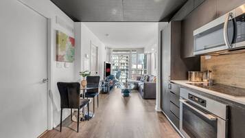 A modern kitchen with a built-in oven and microwave, a dining area with a glass table and chairs, and a living room with a sofa and a TV.