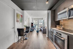Eine moderne Küche mit eingebautem Ofen und Mikrowelle, ein Essbereich mit Glastisch und Stühlen sowie ein Wohnzimmer mit Sofa und Fernseher.