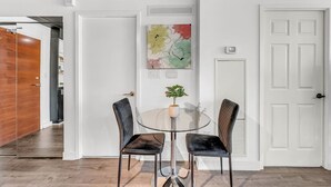 Ein Zimmer mit Glastisch und zwei Stühlen, einer weißen Tür sowie einer Wand mit einem farbenfrohen abstrakten Gemälde.