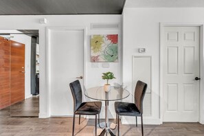 Ein Zimmer mit Glastisch und zwei Stühlen, einer weißen Tür sowie einer Wand mit einem farbenfrohen abstrakten Gemälde.