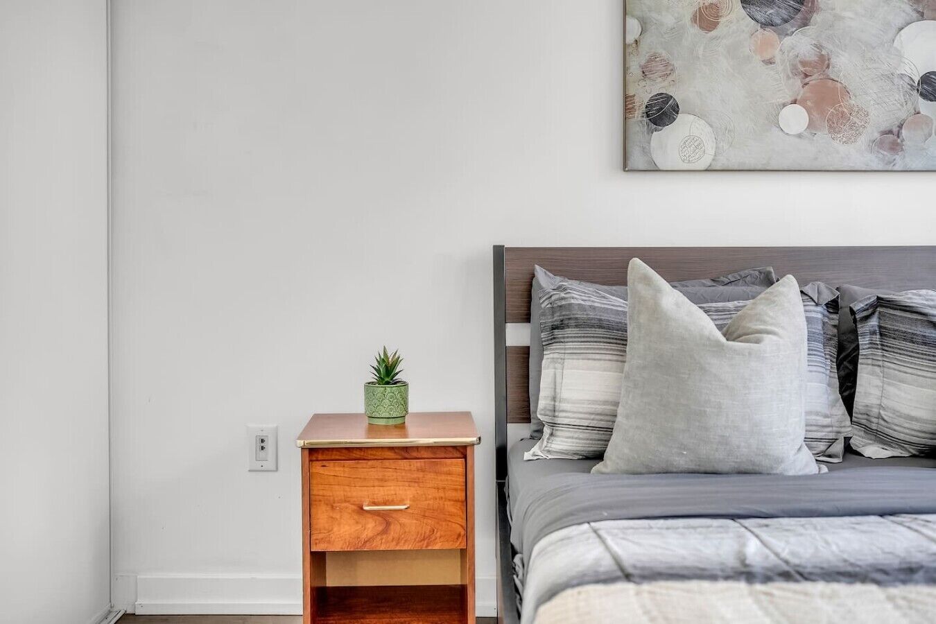 A bedroom with a wooden nightstand, a green plant, a bed with grey bedding and striped pillows, and a wall-mounted abstract painting.