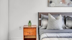 Ein Schlafzimmer mit einem Nachttisch aus Holz, einer grünen Pflanze, einem Bett mit grauer Bettwäsche und gestreiften Kissen sowie einem an der Wand hängenden abstrakten Gemälde.