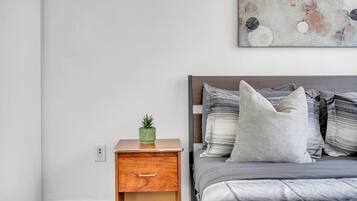 A bedroom with a wooden nightstand, a green plant, a bed with grey bedding and striped pillows, and a wall-mounted abstract painting.