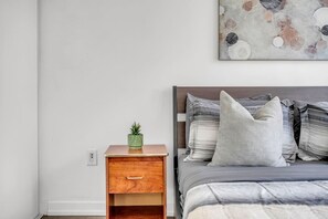 Ein Schlafzimmer mit einem Nachttisch aus Holz, einer grünen Pflanze, einem Bett mit grauer Bettwäsche und gestreiften Kissen sowie einem an der Wand hängenden abstrakten Gemälde.