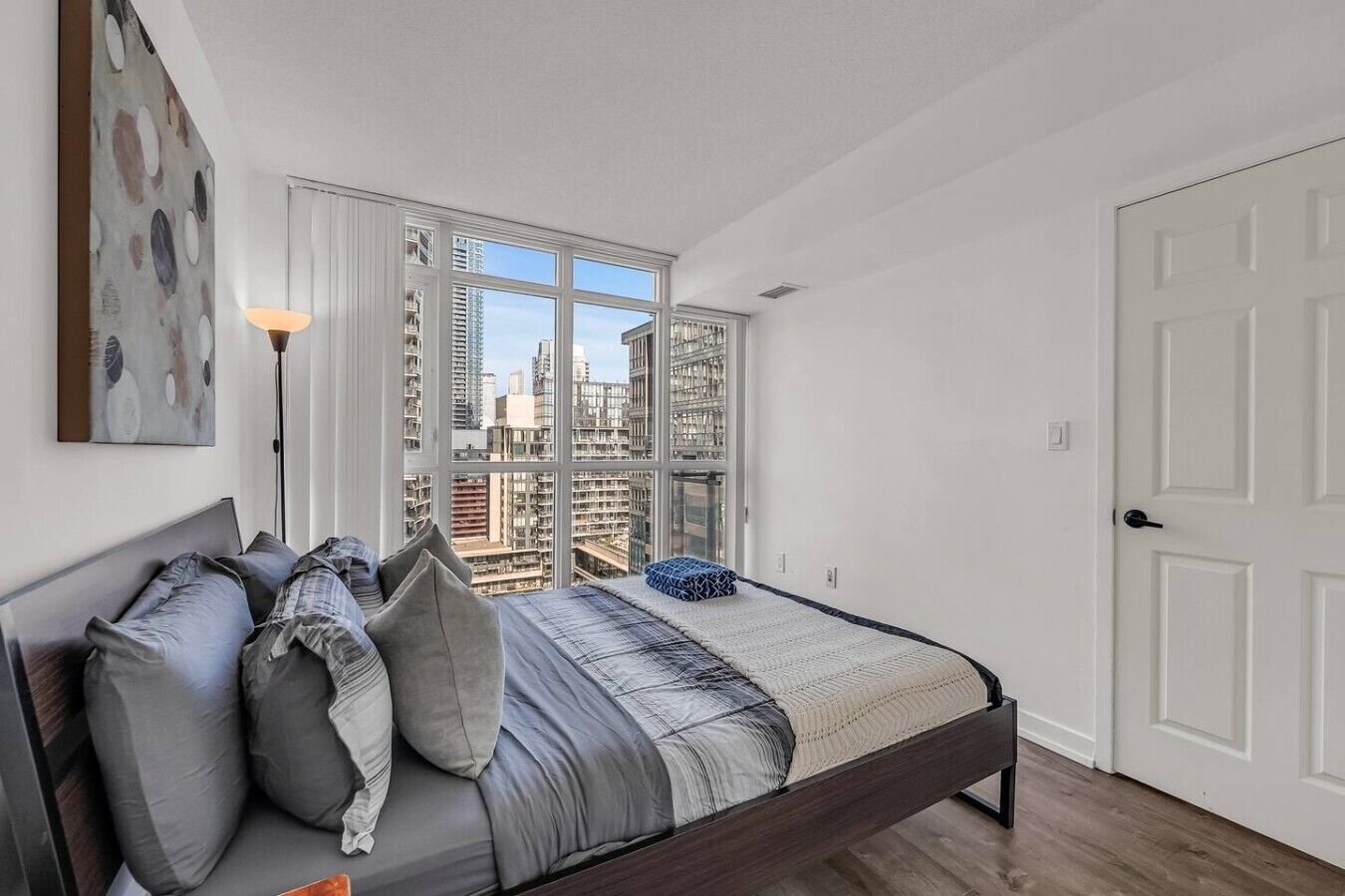 A bedroom with a bed, pillows, a bedside lamp, a view of buildings through large windows, and a door.