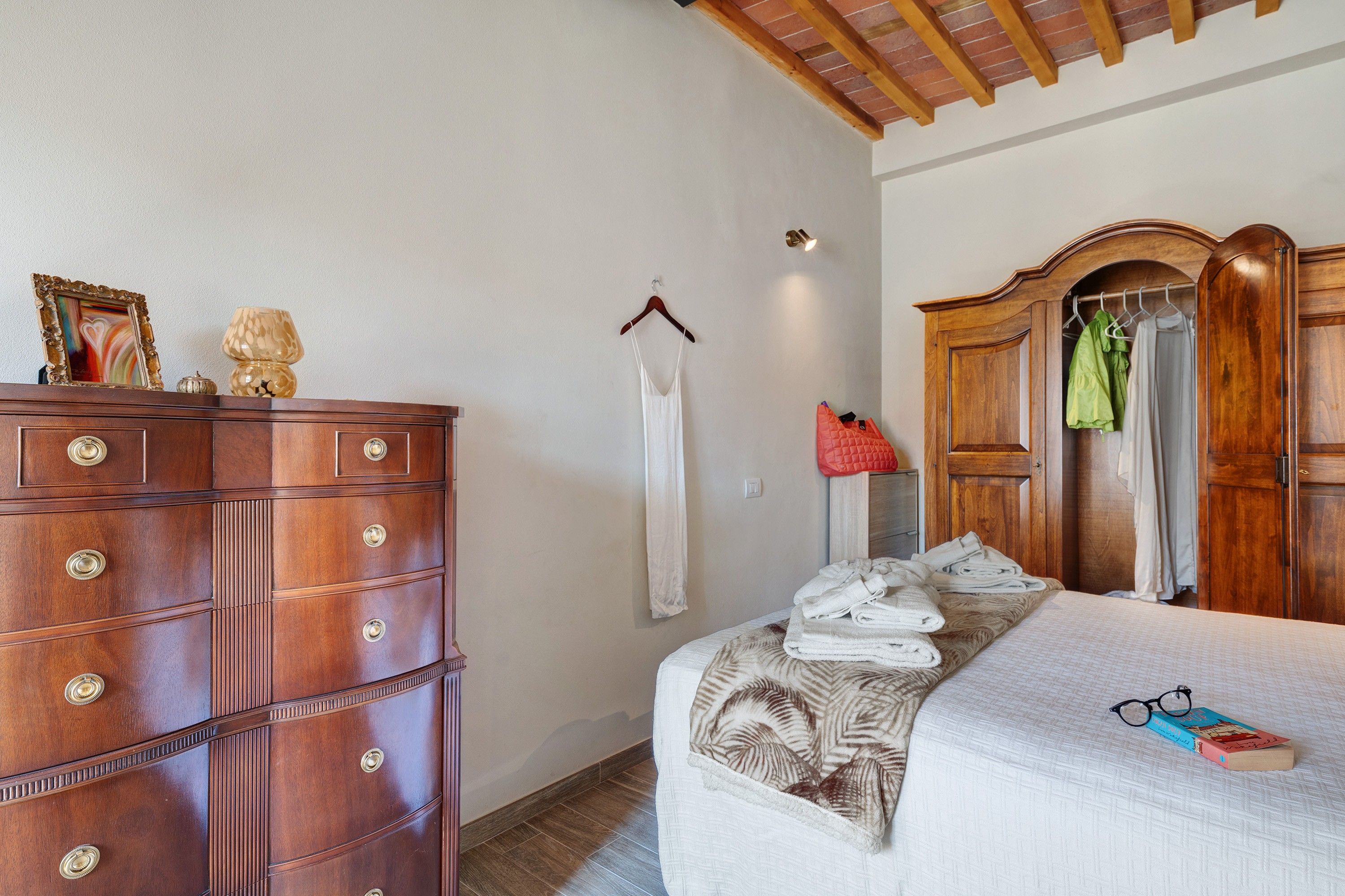 A bedroom with a wooden dresser, a bed with a patterned blanket, and a wardrobe with hanging clothes.