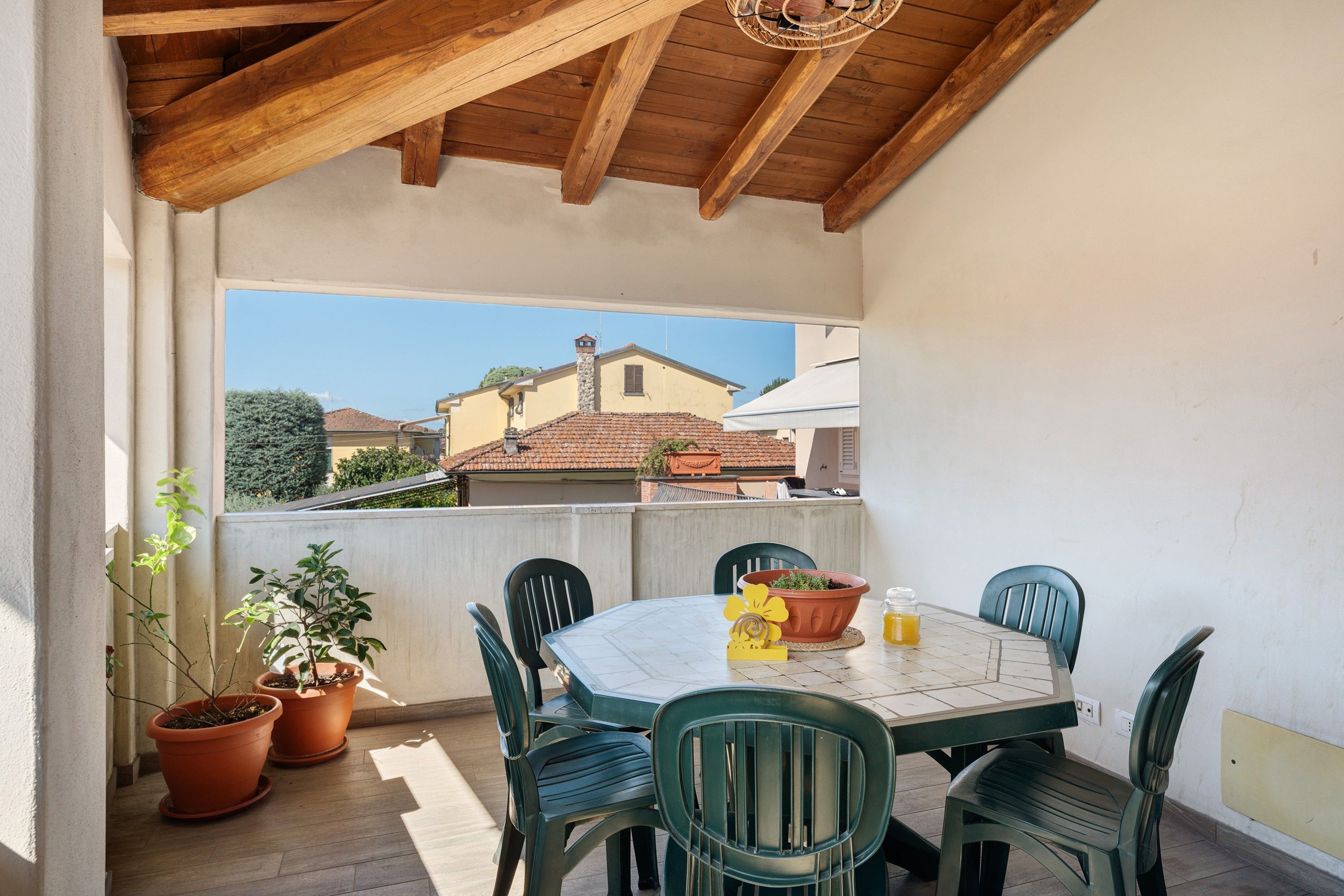 An outdoor patio with a wooden ceiling, a table with chairs, potted plants, and a view of neighboring houses.