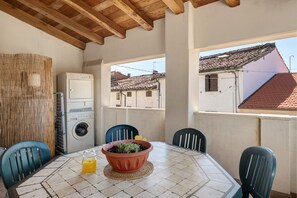 Habitació amb terra de rajola, una taula amb caires, una rentadora i vista a edificis a través d'una finestra.