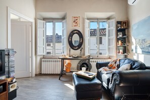 A living room with a black leather sofa, a coffee table, a radiator, a window with a view of buildings, and a shelf with books and decorative items.