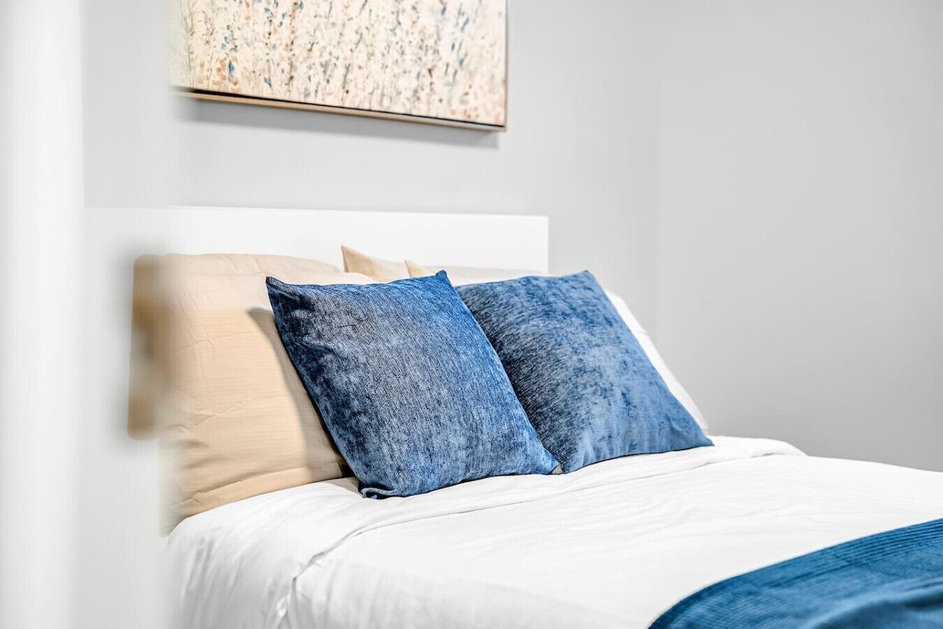 Een bed met witte beddengoed en twee kussens, één beige en één blauw, in een kamer met een effen witte muur en een kunstwerk met textuur erboven.