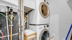 A compact laundry room with a stacked washer and dryer, a wooden shelf with folded towels, and visible plumbing. - *Minutes to BMO Field* 3BR Home in Downtown (Toronto)