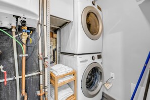 Ein kleiner Waschraum mit übereinander angeordneter Waschmaschine und Trockner, einem Holzregal mit gefalteten Handtüchern und sichtbarer Sanitärinstallation.