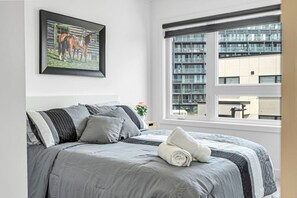 A bedroom with a bed, pillows, a framed picture, a window, and a view of buildings.