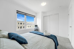 A bedroom with a bed, a window, a door, and a view of buildings outside.