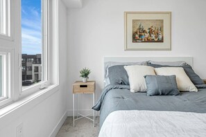 A bedroom with a bed, a bedside table with a plant, a framed picture on the wall, and a window with a view of buildings.