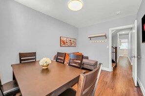 A dining area with a wooden table, chairs, and a vase of flowers. There is a sofa with cushions and a painting on the wall. A hallway leads to another room. - *1 Minute to BMO Field* 4 Bedroom Home In Downtown (Toronto)