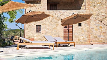 Un espace aménagé au bord de la piscine, avec des chaises longues et des parasols, un bâtiment en pierre doté d’une fenêtre, et une piscine à l’eau d’un bleu limpide.