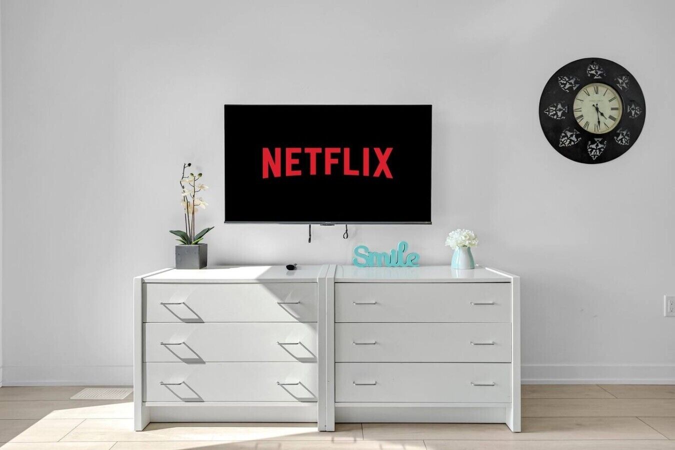A modern living room with a white entertainment unit, a wall-mounted TV displaying 'NETFLIX', a decorative clock, and a 'Smile' sign.