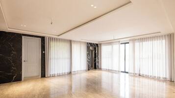 A spacious, modern room with a marble floor, large windows with sheer curtains, and a sleek ceiling design.
