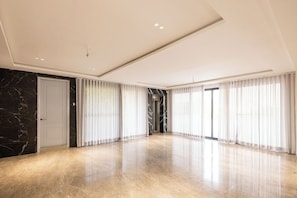 A spacious, modern room with a marble floor, large windows with sheer curtains, and a sleek ceiling design.