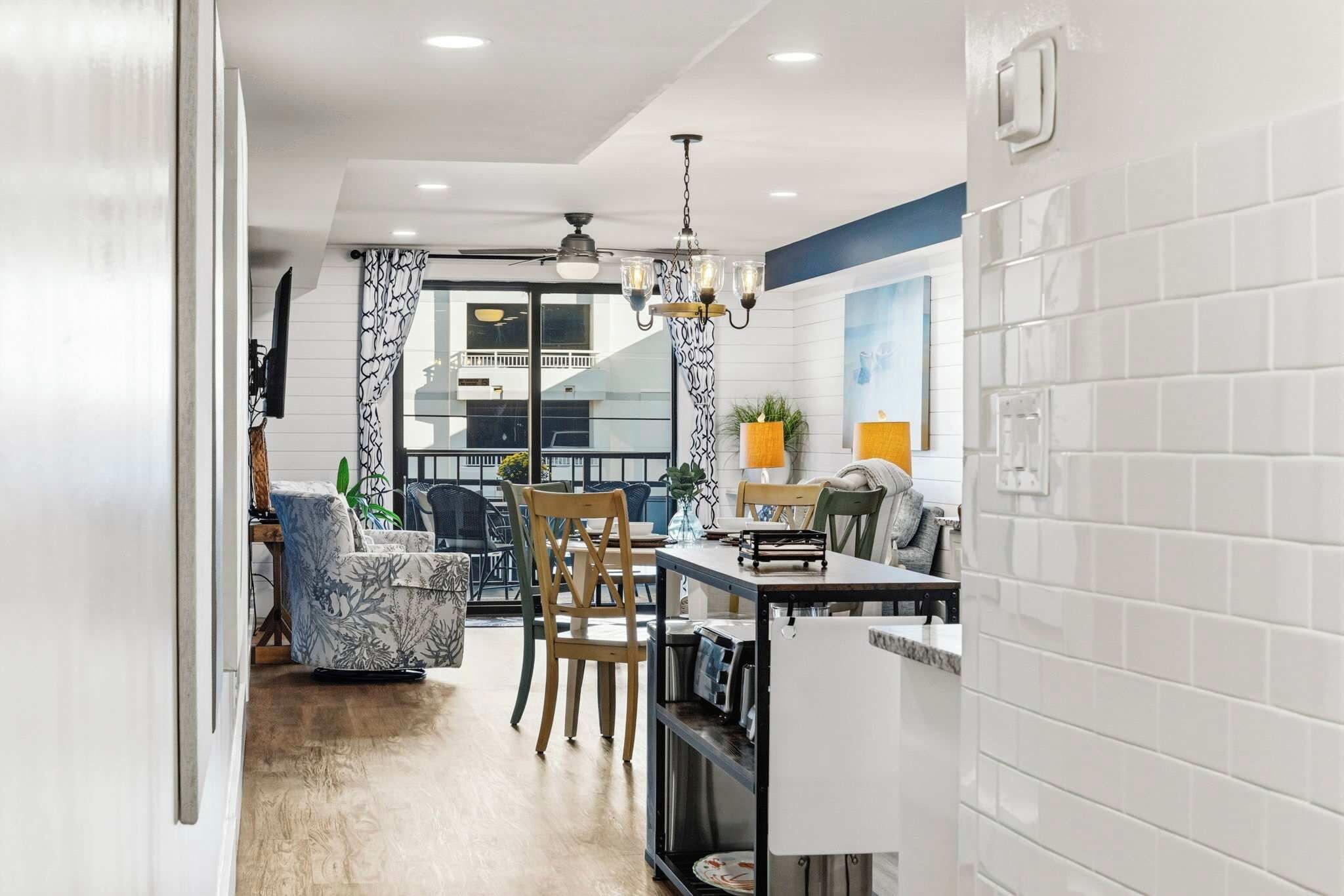 A modern kitchen with a dining area, featuring a chandelier, white tiled walls, and a view of the outside balcony.