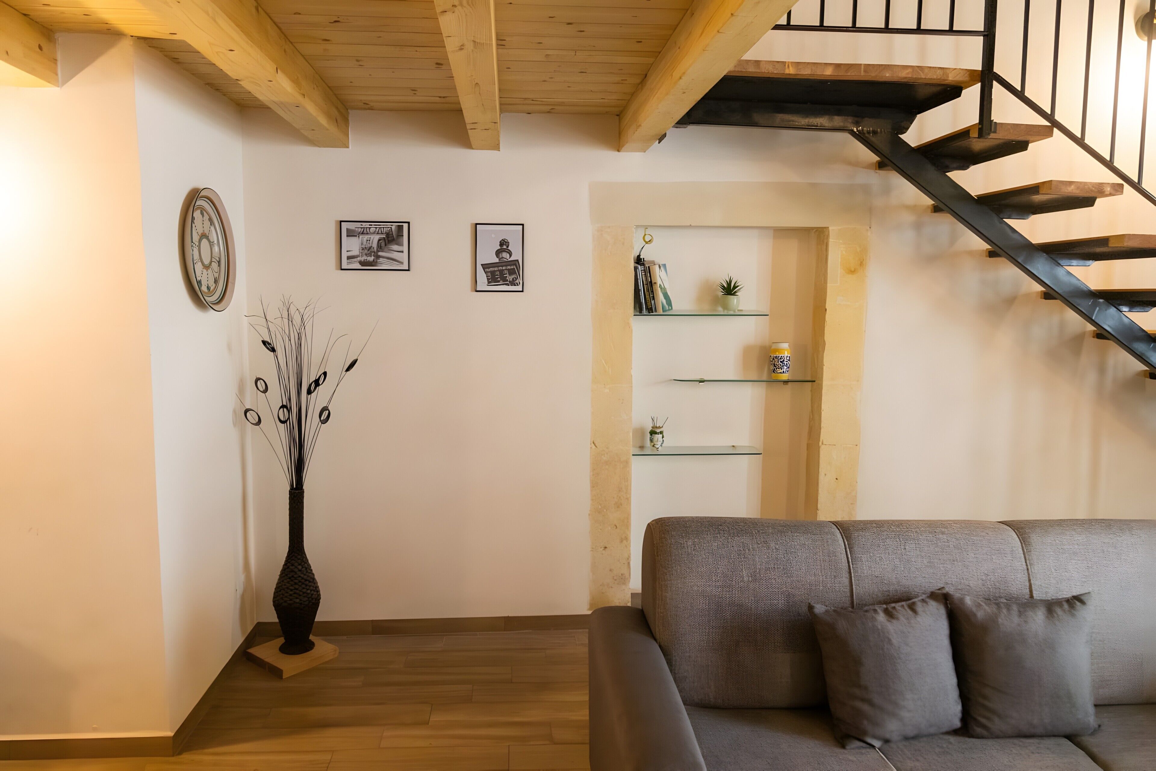 Een woonkamer met een grijze bank, een houten vloer, een trap met een zwarte leuning, en een wand met ingekaderde foto's en een klok.