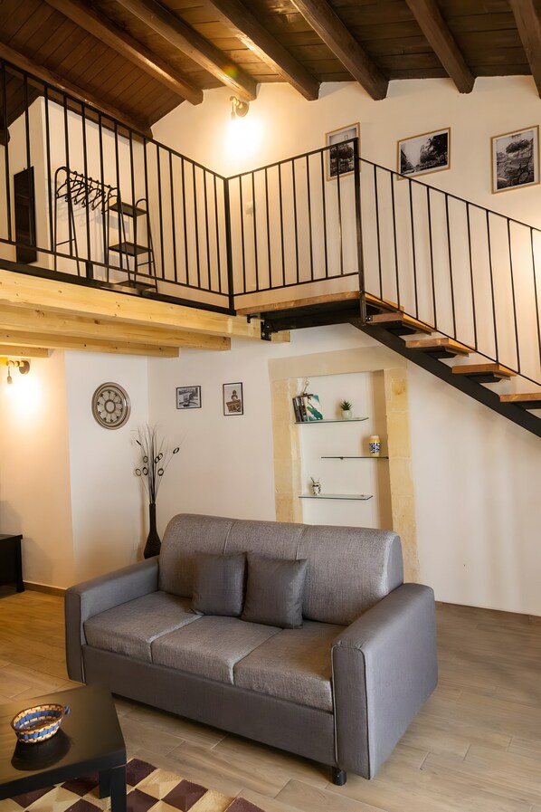 A living room with a grey sofa, wooden ceiling beams, a staircase with a black railing, and wall-mounted shelves.