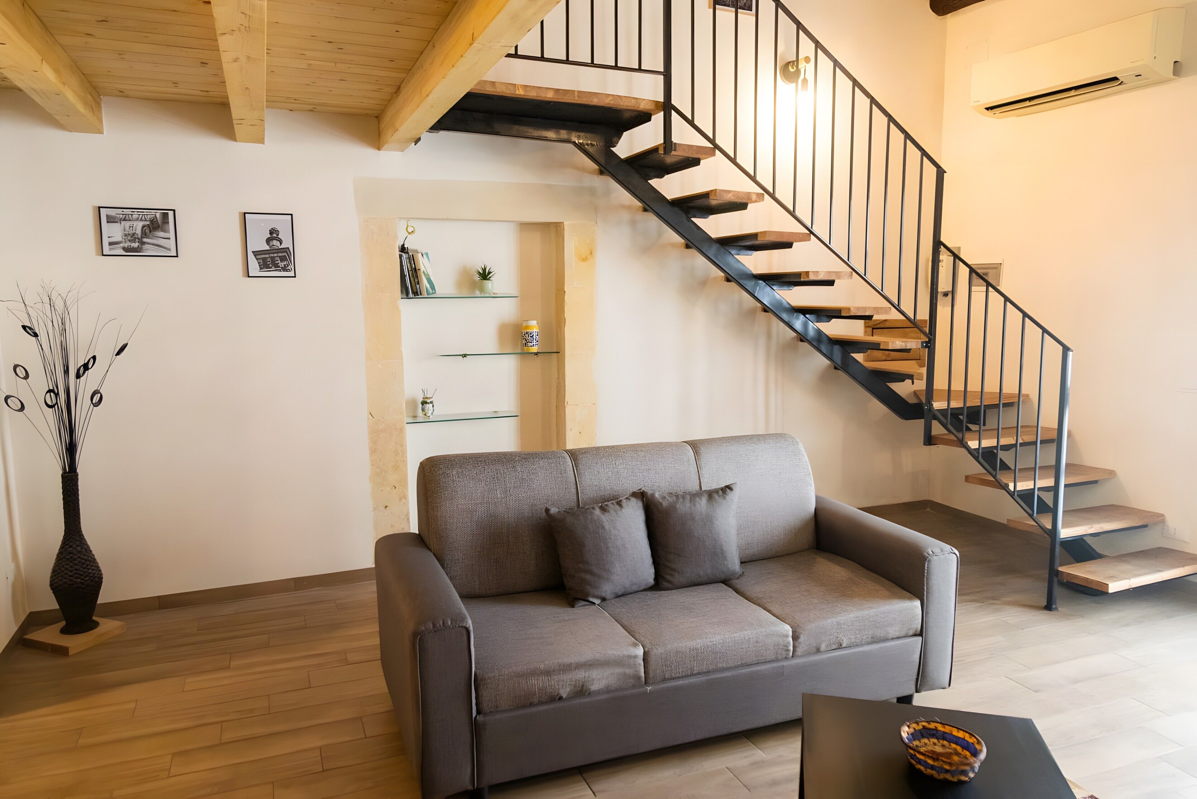 Un salon moderne avec un canapé gris, une table basse en bois, un vase noir, un escalier avec des rampes noires et un mur blanc orné de tableaux.