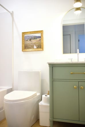 Et badeværelse med toilet, vask, skab, spejl og et billede i ramme på væggen.