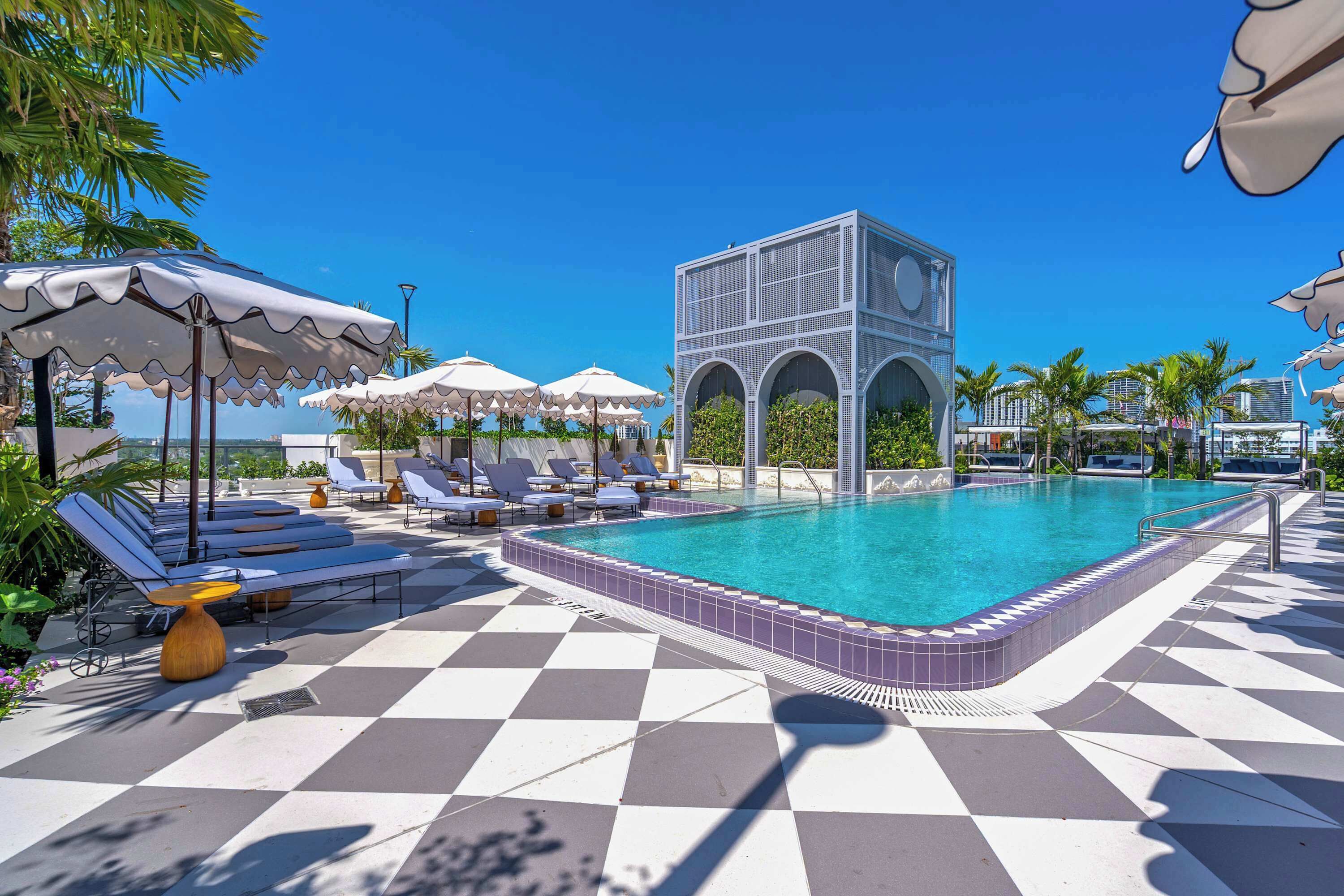 A pool area with lounge chairs and umbrellas, a checkered floor, and a modern building structure.