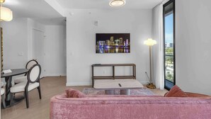 Ein modernes Wohnzimmer mit einem rosafarbenen Sofa, einem Essbereich mit Tisch und Stühlen sowie Blick auf die Stadt durch ein großes Fenster.