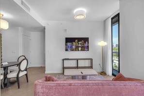 A modern living room with a pink sofa, a dining area with a table and chairs, and a cityscape view through a large window. - Wynwood Luxury Atelier Resort (Miami)