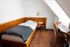 Una habitación pequeña con cama individual, cabecera de madera, mesita de noche con lámpara y una silla.