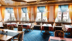 A restaurant with multiple tables and chairs, large windows with curtains, and a patterned carpet.
